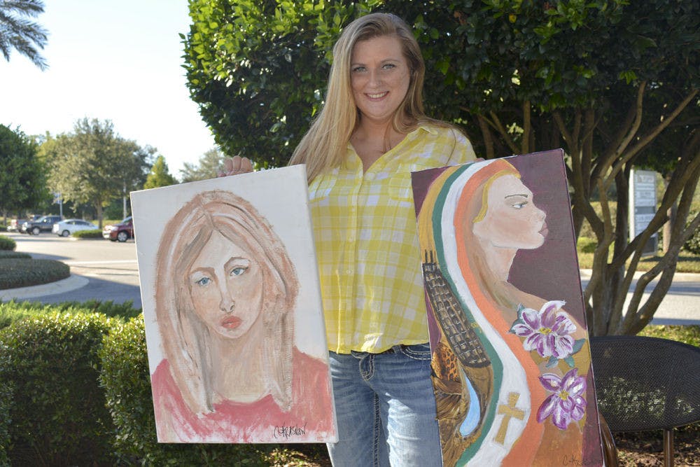 Charissa Krasnow, 35, holds up her two self portraits outside of Starbucks at Tioga Town Center. The left portrait is untitled, and “Warrior” is a mixed-media piece.