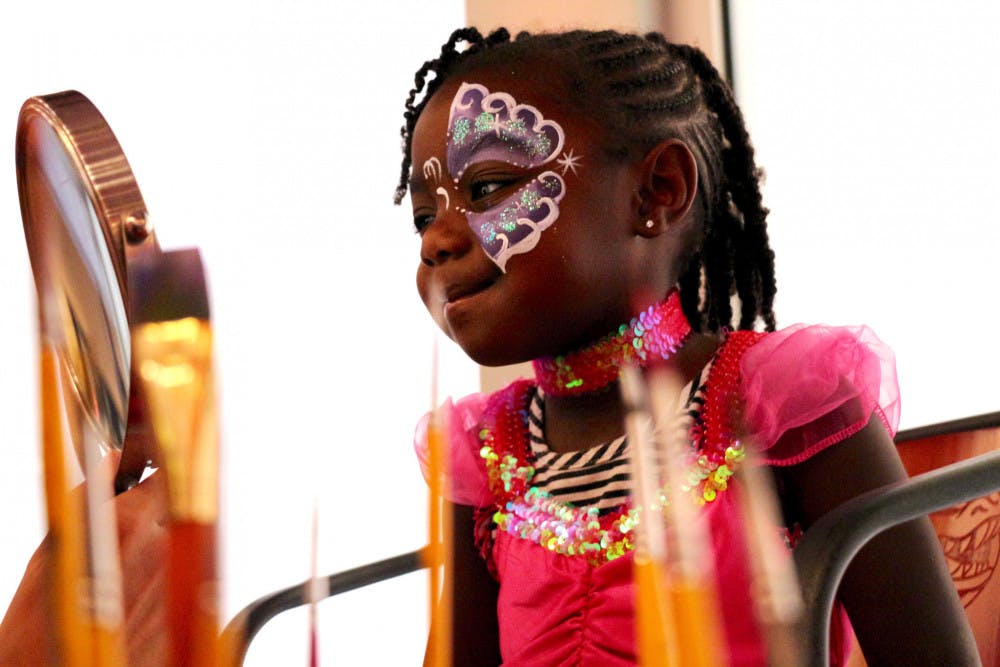 Aulani Newton, 3, a first-timer at Chomp or Treat, looks at her reflection in the mirror after having her face painted as a purple butterfly by one of the Easyjourney Entertainment employees inside the Evans Champions Club at Ben Hill Griffin Stadium on Monday night for Halloween.