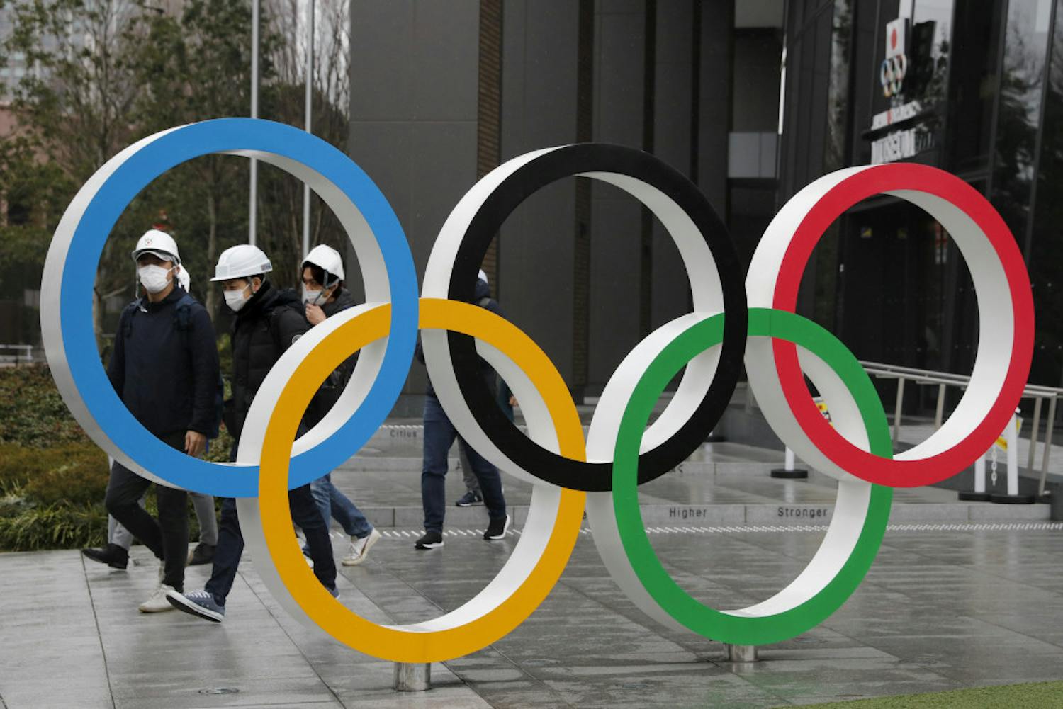 FILE - In this March 4, 2020, file photo, people wearing masks walk past the Olympic rings near the New National Stadium in Tokyo. It's been 2 1/2 months since the Tokyo Olympics were postponed until next year because of the COVID-19 pandemic. So where do the games stand? So far, many ideas about how the Olympic can take place are being floated by the International Olympic Committee, Japanese officials and politicians, and in unsourced Japanese newspaper articles coming from local organizers and politicians. The focus is on soaring costs, fans, or no fans, possible quarantines for athletes, and cutting back to only “the essentials." (AP Photo/Jae C. Hong, File)