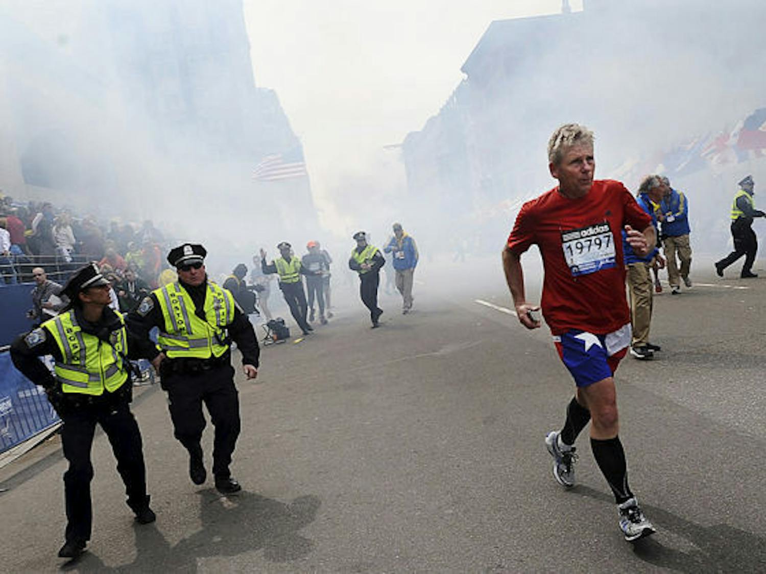 A Boston Marathon competitor and Boston police run from the area of an explosion near the finish line in Boston on Monday. (AP Photo/MetroWest Daily News, Ken McGagh)