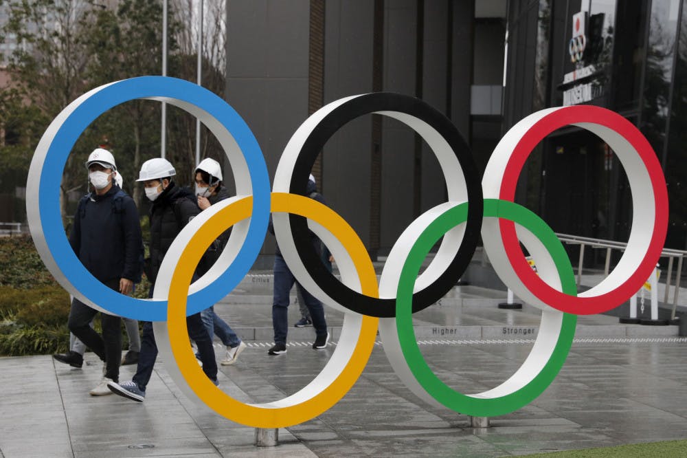 FILE - In this March 4, 2020, file photo, people wearing masks walk past the Olympic rings near the New National Stadium in Tokyo. It's been 2 1/2 months since the Tokyo Olympics were postponed until next year because of the COVID-19 pandemic. So where do the games stand? So far, many ideas about how the Olympic can take place are being floated by the International Olympic Committee, Japanese officials and politicians, and in unsourced Japanese newspaper articles coming from local organizers and politicians. The focus is on soaring costs, fans, or no fans, possible quarantines for athletes, and cutting back to only “the essentials." (AP Photo/Jae C. Hong, File)