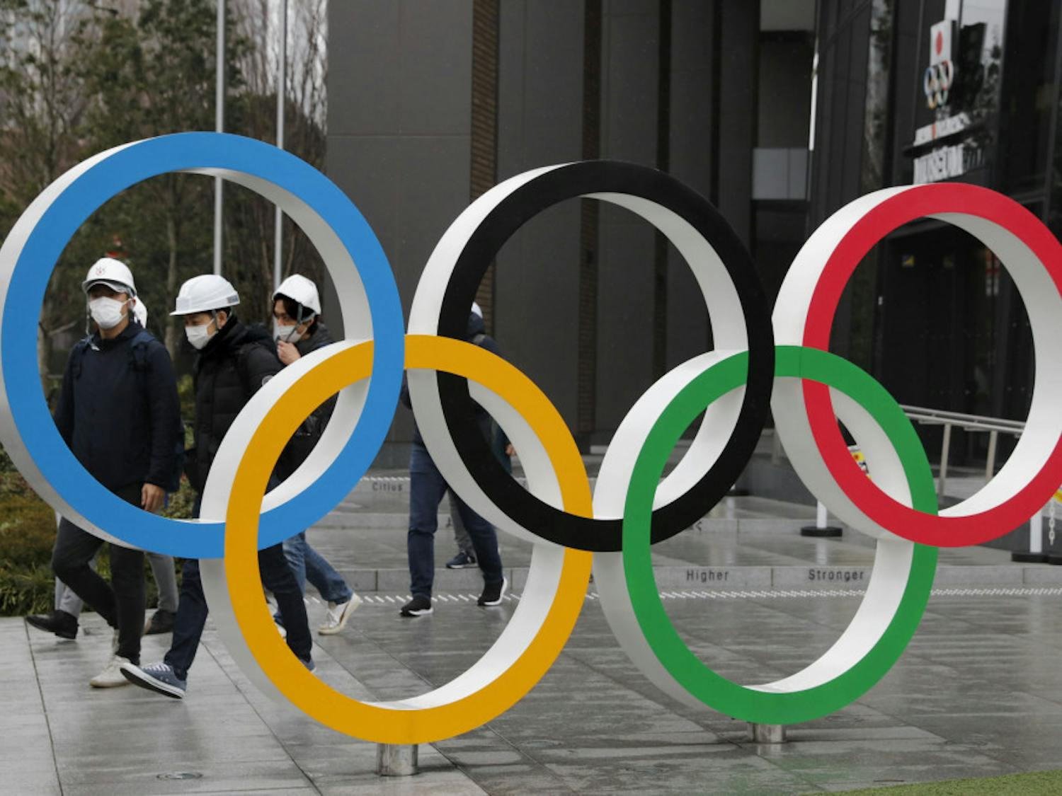 FILE - In this March 4, 2020, file photo, people wearing masks walk past the Olympic rings near the New National Stadium in Tokyo. It's been 2 1/2 months since the Tokyo Olympics were postponed until next year because of the COVID-19 pandemic. So where do the games stand? So far, many ideas about how the Olympic can take place are being floated by the International Olympic Committee, Japanese officials and politicians, and in unsourced Japanese newspaper articles coming from local organizers and politicians. The focus is on soaring costs, fans, or no fans, possible quarantines for athletes, and cutting back to only “the essentials." (AP Photo/Jae C. Hong, File)